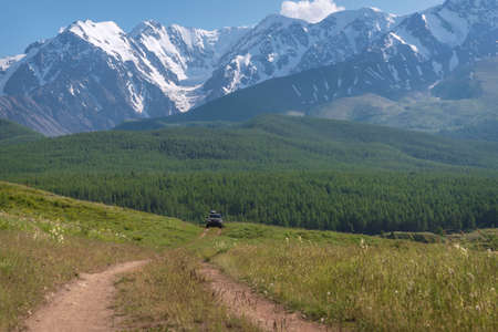 Amazing summer view with snowy mountains, glacier, green grass, dirt road and car against blue sky and clouds. Altai, Russiaの写真素材