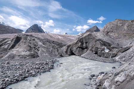 Amazing view of a glacier with cracks, snow, ice and a river with white water flowing from the glacier against the backdrop of snowy peaks and a blue sky with clouds. Altai, Russiaの写真素材
