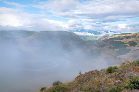 Amazing morning summer landscape with mountains covered with snow and forest, meandering river, asphalt road and fog against blue sky and cloudsの写真素材