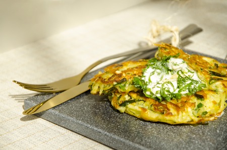Roesti. Potatoes grated with greens, fried in butter. Serve on a stone board with sour cream, garlic and greens. Served with textile napkin and cutlery. Vegetarian table. Horizontal. Copy space.の写真素材