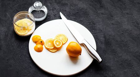 Dark moody. Food black background. On a white stone board in the hands of a girl, a knife cuts a lemon. Slices of lemon are piled in a glass jar and sprinkled with sugar. In the foreground is a whole lemon. Close-up. Copy space.の写真素材