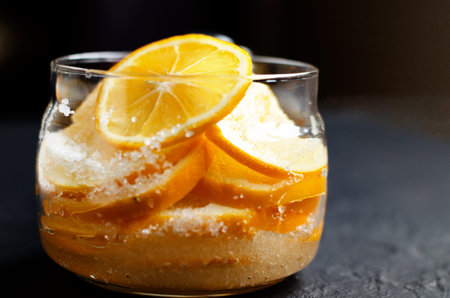 Dark moody. Food black background. Slices of lemon are piled in a glass jar and sprinkled with...