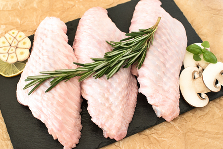 raw parts of turkey wing, three pieces of chicken, on a round wooden cutting board with mushrooms and rosemary on a black stone desk and brown paper backgroundの写真素材