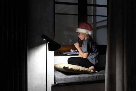 Boy wearing a santa claus hat writes a letter asking for a gift for christmas in a dark room