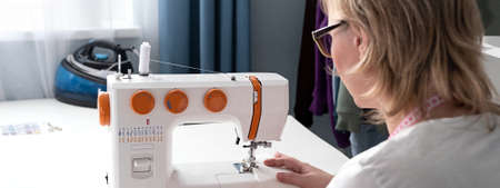 Real middle aged woman tailor sewing clothes using a sewing machine at home. Seamstress working. Hand making clothes in home interior. Female sits enjoying the process of sewing clothes. creative skills design, hobbies, lifestyle and small business concept.の写真素材