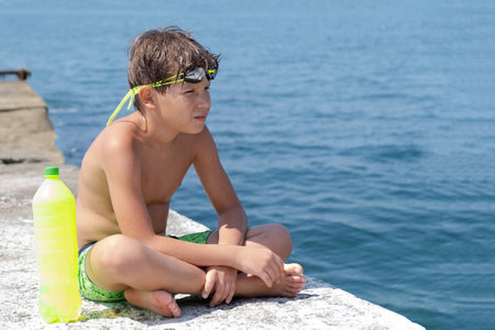 A cute boy in swimming glasses and shorts with a bottle of water sits on a pier on the seashoreの写真素材