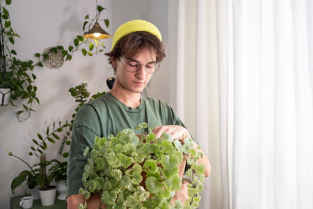 A young handsome man with dark long hair, wearing a green T-shirt, hat and glasses holds a houseplant in his hands.の写真素材