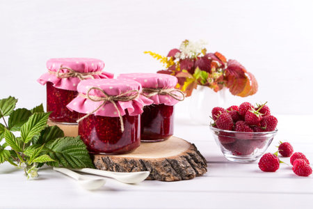 Raspberry jam in glass jar, fresh  ripe raspberry and green leaves on white wooden table