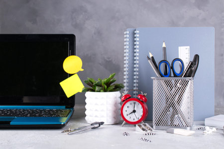 Laptop, red alarm clock and supplies on a gray concrete desk. Back to school concept.