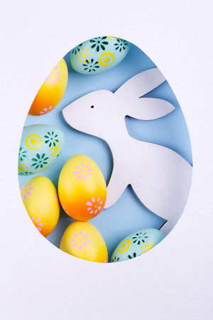 Easter egg frame made of paper, colorful painted chicken eggs, white wooden bunny on blue background. Easter composition. Flat lay, top view, copy spaceの写真素材