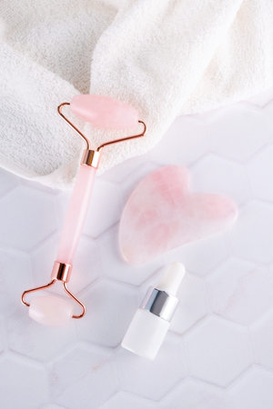 Pink face roller and gua sha massager made from natural quartz stone with cosmetic massage oil over white background. Lifting and toning treatment at home.の写真素材