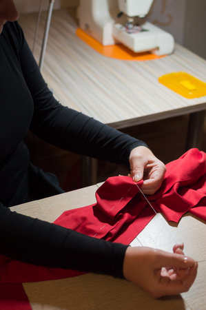 Dressmaker's hands sewing red fabric with needle and thread on table in sunlight