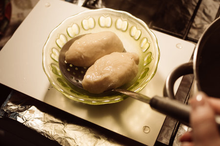 Putting hot fresh cottage cheese dumplings with slotted spoon from aluminum saucepan into plate on stove in kitchenの写真素材