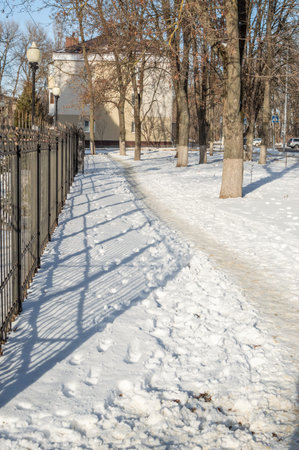 A path, footprints in the snow and the shadow of an iron fence on a sunny winter afternoon in a city parkの写真素材