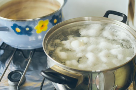 Lots of white fresh organic chicken eggs are boiled in boiling water with foam in a saucepan on the stoveの写真素材