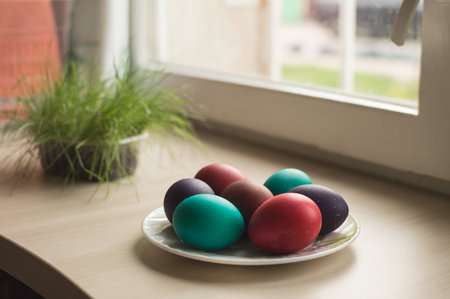 Seven colorful Easter eggs on a plate and green grass on a light beige table by the windowの写真素材
