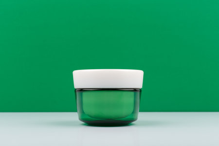 Green glass cream jar with white cap on white table against green background with space for textの写真素材