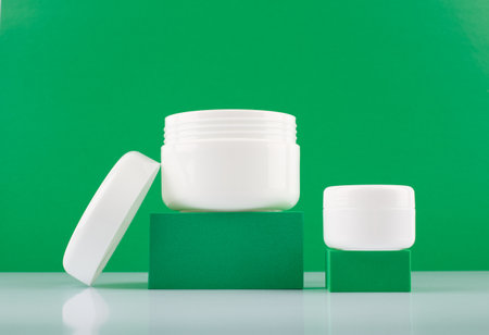 Close up of two white glossy cream jars on podiums against green background with space for text. Concept of beautyの写真素材