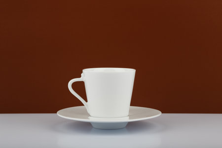 White shiny coffee cup on a plate on white table against brown background with copy spaceの写真素材