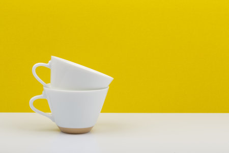 Two white coffee cups on in another on white table against yellow background with copy spaceの写真素材