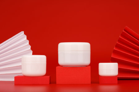 Cream jars on red podiums against red background decorated with wavers. Concept of luxury skin care