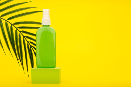 Spray for sun protection with sunscreen in green tube on podium against yellow background with palm...