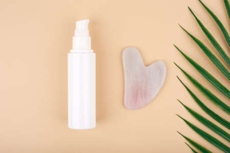 Top view of skin lotion, rose quartz heart shaped guasha massage stone and half of palm leaf on light beige background.の写真素材
