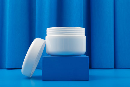 White opened cosmetic jar on blue pedestal against blue fabric background with waves. Concept of luxury skin care products, face cream or mask for glowing, young looking skinの写真素材