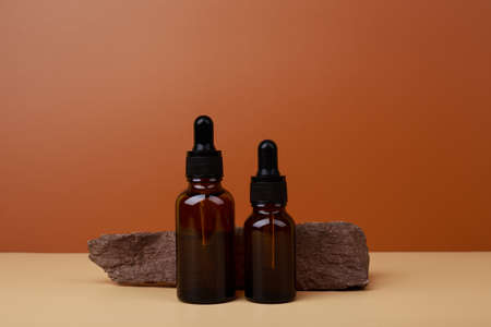 Two dark glass bottles with serum or oil for skin care next to stone on beige table against brown background. Cosmetic bottles with natural rough stone against dark backgroundの写真素材