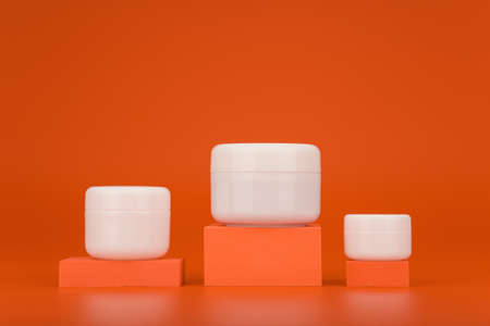 Set of three cosmetic jars of different sizes on podiums against orange background with copy space. Concept of cream, lotion, mask or scrub with vitamin C for anti aging or regular skin careの写真素材
