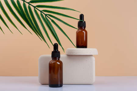 Two dark transparent glass bottles with skin serum or oil for manicure on gypsum podium against beige background with palm leafの写真素材