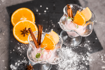 Ice cream dessert, yogurt with orange slices, green mint leaves, cinnamon sticks on a dark stone tray and gray concrete background.の写真素材