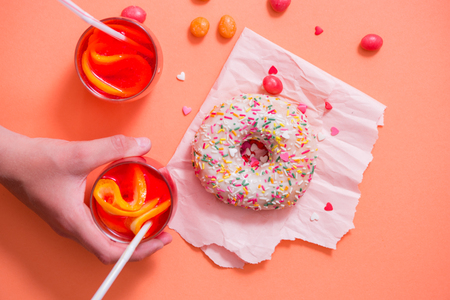 Child's hand takes sweet pastry. Frosted sprinkled donut on pink background. Tubules for a cocktail. Spilled orange candy. Cold drink from lemon orange and lime with ice in a glass cupの写真素材