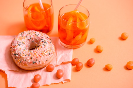 Child's hand takes sweet pastryFrosted sprinkled donut on pink background. Tubules for a cocktail. Spilled orange candy. Cold drink from lemon orange and lime with ice in a glass cupの写真素材