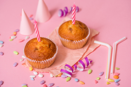 two chocolate muffins, a birthday candle. Party for girls. Caps and tinsel and multi-colored sweets on a pink backgroundの写真素材