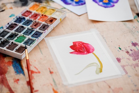 Watercolor paints, brushes and drawing paper on a white table by the window next to glasses, flower vases and a lamp. Top view, flat layの写真素材