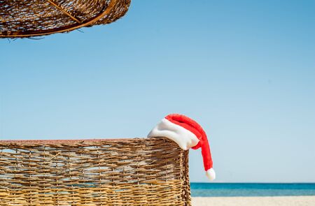Santa's hat on a tropical beach.Blue sky and white sand, Egyptian or Turkish beach and sea. Winter holidays, hot sun. Wicker umbrella and deck chair. Christmas on the island.の写真素材