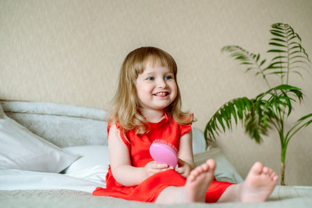 Cute red-haired blue-eyed emotional baby girl laughs. Lies on the bed in the bedroom, the bed is a white bed, a red dress for a child. pillows and blanket. smile and joyの写真素材