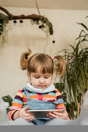 Cute baby girl playing at home on the floor in the phone, face mask, quarantine from the virus covid-19.concept of modern children and Internet in life. life style, exotic indoor plants..の写真素材