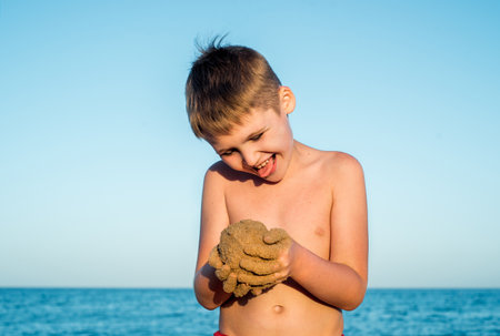 Sea holidays. Cute boy on the beach by the sea plays with sand. Casts a sand castle and cakes. sunset, blue sky and relax travel concept. copy spaceの写真素材