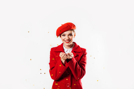 beautiful girl in a red coat and a red beret blowing magic stars. holiday, flying sparkles and tinsel. Birthday party for valentines day or christmasの写真素材