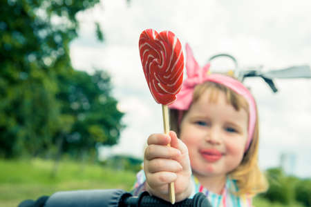 Fashionable baby girl, sunglasses, lollipop like a heart. Happy children hold candy green grass outdoors. Candy shop. Lollipop or sucker on sticks. Sugary treats. Sweet dreamの写真素材