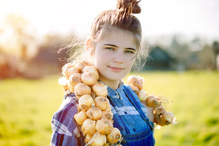 Happy Teenager girl in summer yellow boxing field. Girl farmer preparing for planting bulbs. Bouquet of onions in the hands. Sunny warm day in the villageの写真素材