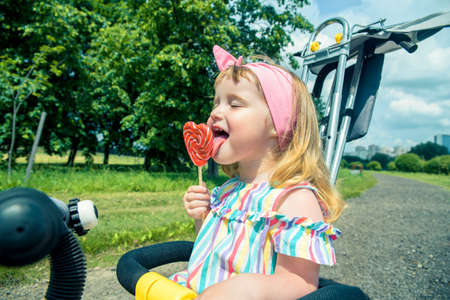 Cute curly baby girl eating watermelon candy in a sunny park. baby with outdoors lollipopの写真素材