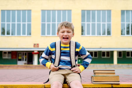 sad displeased schoolboy, the boy cries and does not want to go to school. outdoors at the day time. Sitting on the steps with books and a large school backpack.Concept school themeの写真素材