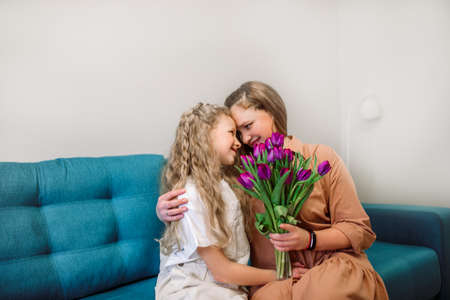Cute little girl greeting her mother at home. Bouquet of purple tulips. Mothers day conceptの写真素材