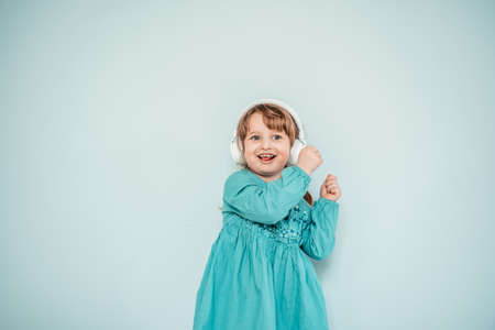 caucasian baby girl in white headphones listens to music and dances in a turquoise blue bright dressの写真素材