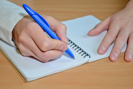 A man's hand in a white shirt writes the text with a blue pen in a notebook with a spiral on a wooden tableの写真素材