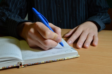 A man's hand in a blue shirt writes the text with a black pen in a notebook on a wooden table, with...