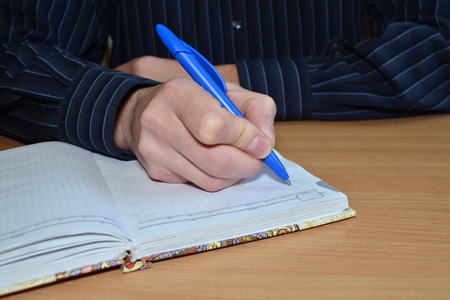 A man's hand in a blue shirt writes the text with a blue pen in a notebook on a wooden tableの写真素材